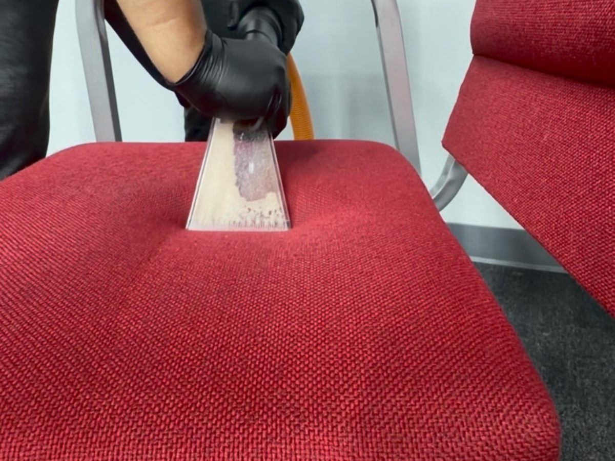 Gloved hand running an upholstery extraction tool across a red chair seat, with soil visible inside the clear head